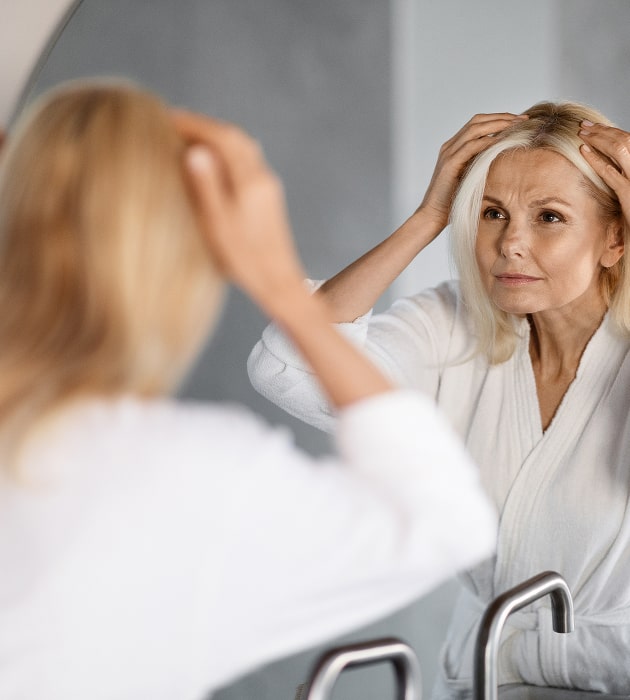 Mature woman in white robe examining scalp and hairline in mirror, showing thinning hair concerns