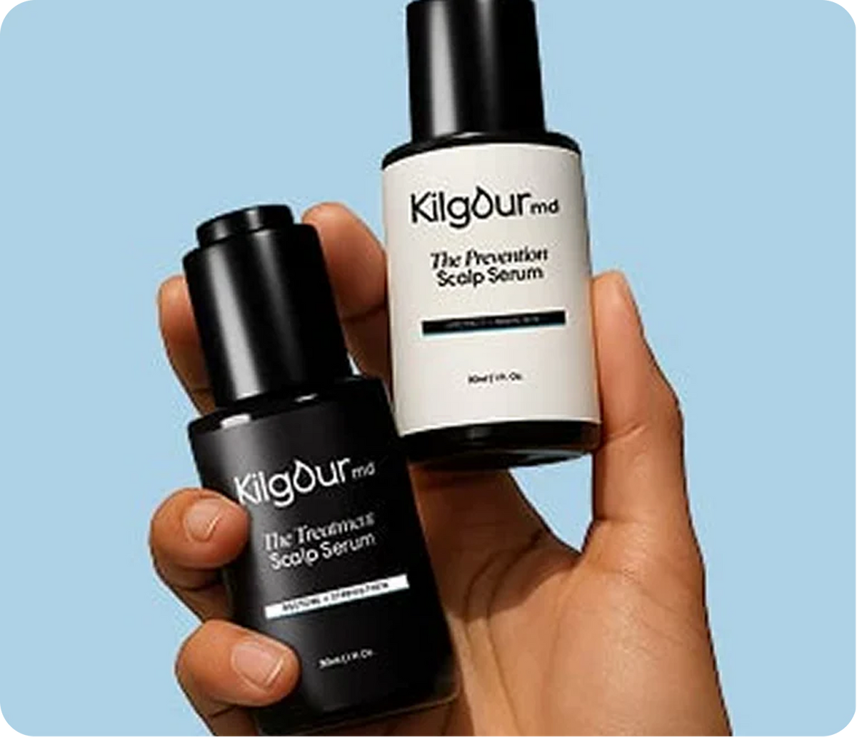 Hand holding two KilgourMD scalp serum bottles (black Treatment Scalp Serum, white Prevention Scalp Serum, 30 mL) against a light blue background
