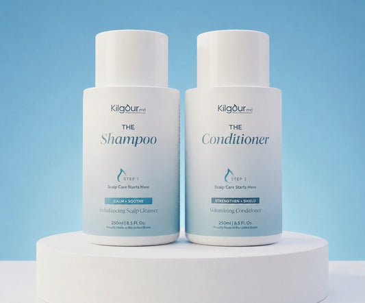 Front view of The Shampoo and Conditioner Bundle: two white KilgourMD bottles labeled The Shampoo and The Conditioner on a white pedestal against a light blue background with a ‘Developed by a Stanford-trained dermatologist’ banner