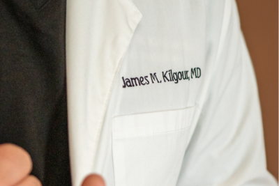 Close-up of a dermatologist in a white lab coat showing the embroidered name on the chest against a warm brown background
