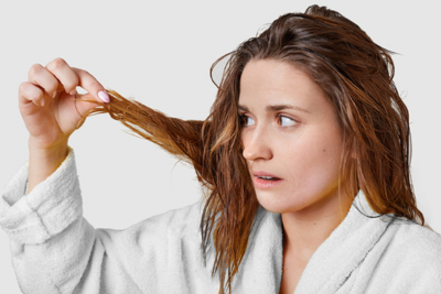 Woman in white bathrobe inspecting wet hair strands with a concerned expression against a pale gray background
