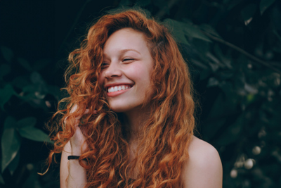 Smiling young woman with long curly red hair against dark leafy background