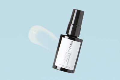 Angled view of The Science of The Prevention serum bottle showcasing a black glass pump bottle and a clear serum smear on a light blue background