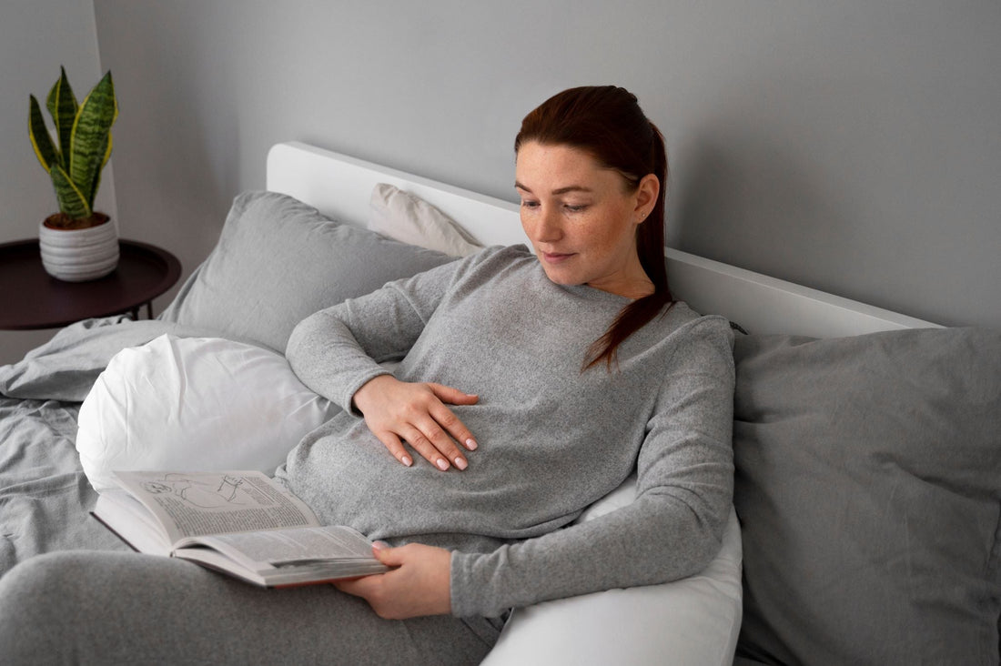 Pregnant woman with brown hair in gray loungewear reclining on bed, hand on belly while reading a book