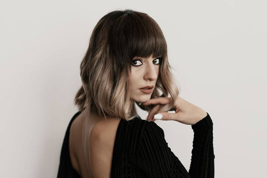 Woman with shoulder-length brown-to-ash-blonde balayage bob and blunt bangs looking over her shoulder, black backless top