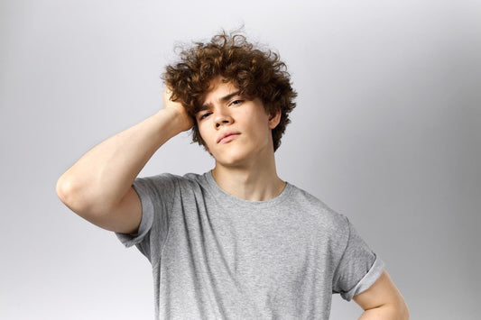 Young man with curly brown hair touching scalp, wearing gray t-shirt — focus on hair and scalp