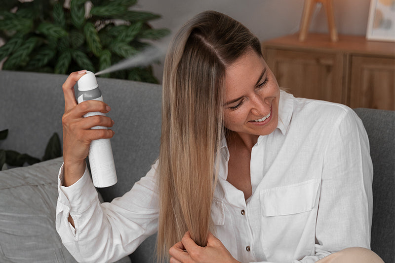 Woman spraying hairspray onto long hair while smiling on a gray sofa in a cozy living room